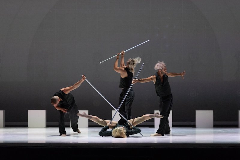 Dancers in vests and slacks hold metal poles. One dancer lays on their back with their legs splayed. The others stand in mid-movement.